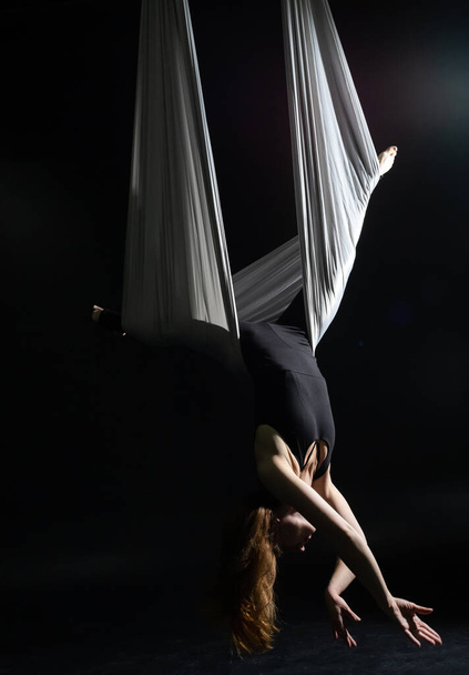 Fly yoga. Girl in a white hammock on a black background shows aerial acrobatics. Gymnastics, circus, under dome. Play of light and shadow creates feeling of a theatrical performance. Sports activity - Photo, Image