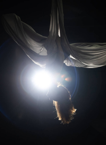 Leć na jogę. Dziewczyna w białym hamaku na czarnym tle pokazuje akrobacje lotnicze. Gimnastyka, cyrk, pod kopułą. Gra światła i cienia tworzy poczucie spektaklu teatralnego. Działalność sportowa - Zdjęcie, obraz