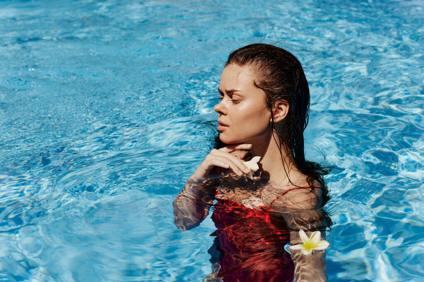 Młoda kobieta w basenie w czerwonym stroju kąpielowym z pięknym uśmiechem i balijski kwiat w słońcu pływanie w basenie, koncepcja relaksu na wakacjach. Wysokiej jakości zdjęcie - Zdjęcie, obraz