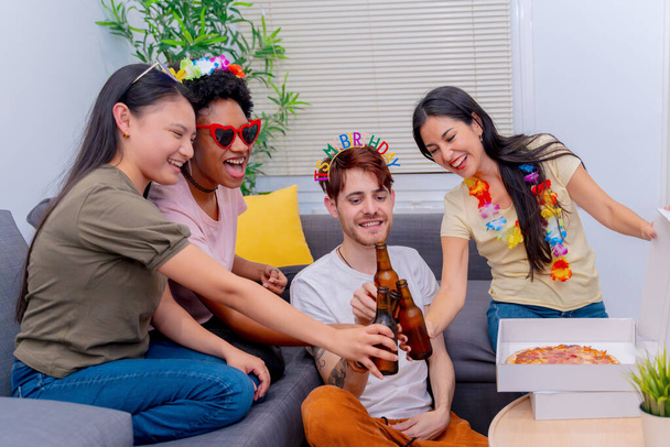 Un grupo jovial de amigos disfrutan de una celebración de cumpleaños con cervezas frías y una pizza fresca en una acogedora sala de estar.  - Foto, Imagen