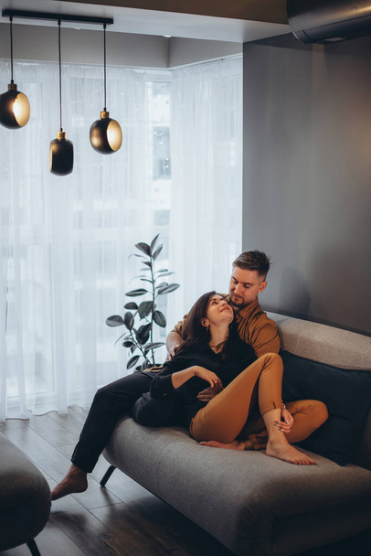 A married young couple sits on a dark sofa in a cozy apartment against the backdrop of a graphite wall and window. Couple hugging and kissing - Photo, Image