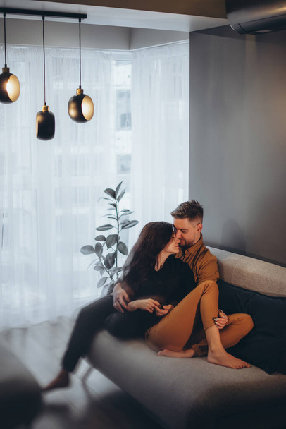 Małżeństwo siedzi na ciemnej sofie w przytulnym mieszkaniu na tle grafitowej ściany i okna. Para przytulająca i całująca - Zdjęcie, obraz