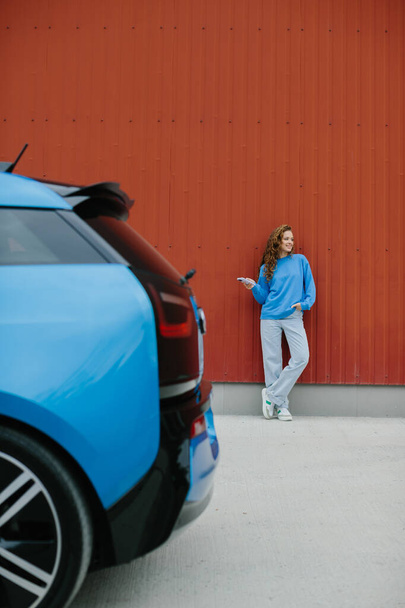 Μια κομψή νεαρή γυναίκα στέκεται δίπλα στο ηλεκτρικό της αυτοκίνητο στο πάρκινγκ.. - Φωτογραφία, εικόνα