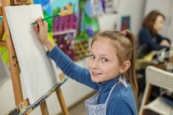 Vidám gyermek lófarok festékek vászonra, élvezi a művészeti osztály körül színes műalkotás a háttérben. - Fotó, kép