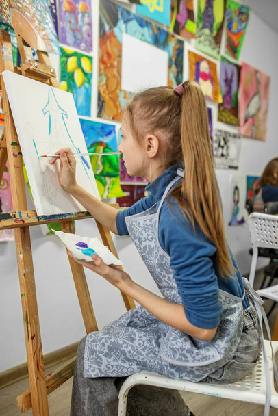 Skupiona młoda dziewczyna w klasie plastycznej maluje pędzlem na białym płótnie, otoczona kolorowymi dziełami sztuki. - Zdjęcie, obraz