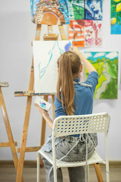 Gericht jong meisje in een kunstklas verf met borstel op wit doek, omgeven door kleurrijke kunstwerken. - Foto, afbeelding