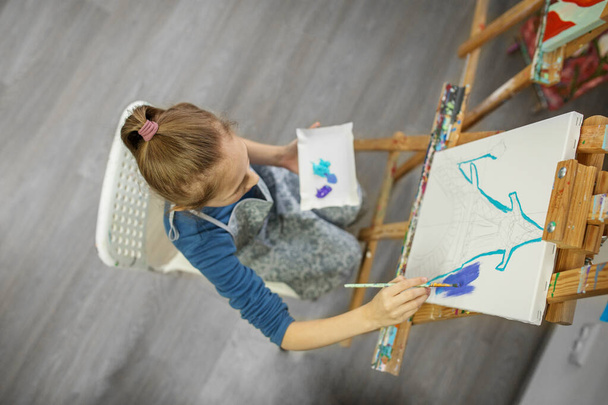 Keskittynyt nuori tyttö, jolla on korkea poninhäntä, imeytyy maalaukseen harjalla maalaustelineessä, mikä osoittaa hänen luovuutensa ja keskittymisensä.. - Valokuva, kuva