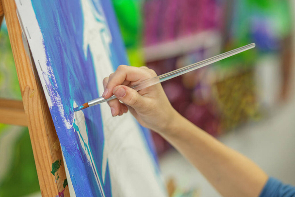 Focused artist's hand paints broad strokes of vivid blue on canvas, detailing motion and technique of painting - Photo, Image