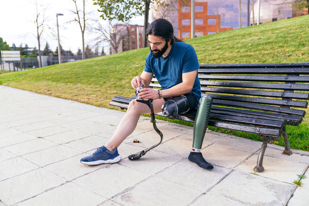 Uomo concentrato con la barba su una panchina del parco, che prepara la gamba protesica per una corsa, mostrando la routine di un atleta adattivo. - Foto, immagini