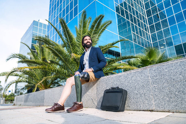 Веселый мужчина-предприниматель с протезной ногой сидит рядом с современным офисом, пальмы на заднем плане, выглядит оптимистично. - Фото, изображение