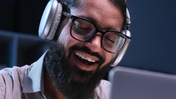 Κοντινό πορτρέτο του Ινδού επιχειρηματία που φοράει ακουστικά, εργάζεται, ακούει μουσική και τραγουδά κατά τη διάρκεια της νυχτερινής εργασίας με φορητό υπολογιστή στο γραφείο. - Πλάνα, βίντεο