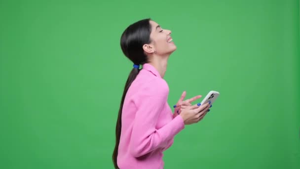 Mujer joven caucásica navegando por Internet, mensajería, mensajes de texto, comprometida en el entretenimiento en línea usando el teléfono, riéndose sobre el fondo verde. - Metraje, vídeo