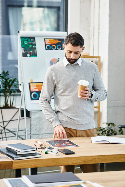 Een man ontspant zich aan een tafel met een kop koffie in een bruisende kantooromgeving, neemt een moment om te hergroeperen te midden van de bedrijvigheid. - Foto, afbeelding