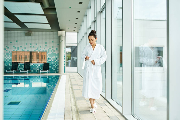 Μια νεαρή, όμορφη μελαχρινή γυναίκα με μπουρνούζι στέκεται δίπλα σε μια εσωτερική πισίνα.. - Φωτογραφία, εικόνα