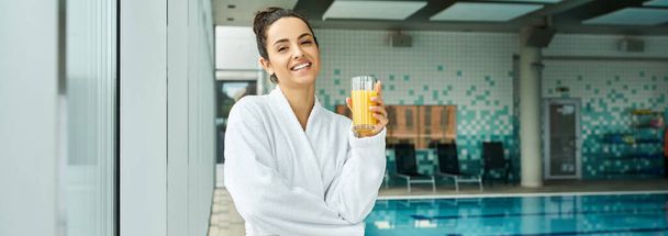 Een brunette in een luxe badjas nipt rustig sinaasappelsap bij een overdekte spa met een zwembad. - Foto, afbeelding