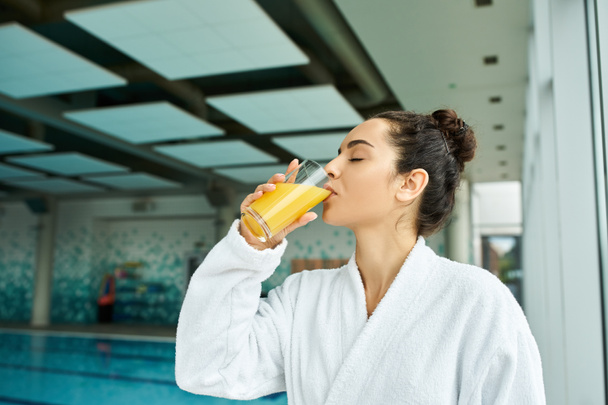 Uma jovem, bela mulher morena saboreando luxuosamente um refrescante copo de suco de laranja em um spa interno por uma piscina. - Foto, Imagem