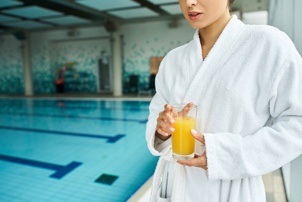 Eine junge, schöne brünette Frau im Bademantel entspannt am Pool und hält ein Glas Orangensaft in der Hand. - Foto, Bild