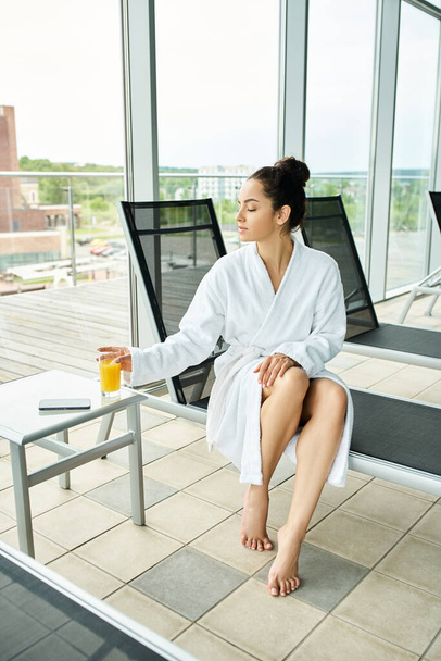 A young brunette woman sits in a bathrobe holding a glass of orange juice. - Photo, Image