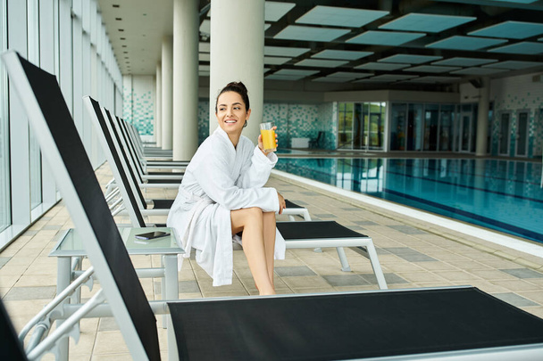 Una giovane donna bruna si siede su una sedia a sdraio vicino a una piscina coperta, tenendo in mano un drink. - Foto, immagini