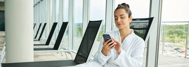 Молодая брюнетка, сидящая в кресле, очарованная экраном мобильного телефона, в крытом спа-салоне с бассейном. - Фото, изображение