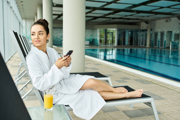 Een jonge, mooie brunette in een badjas zit op een ligstoel bij een overdekt zwembad. - Foto, afbeelding