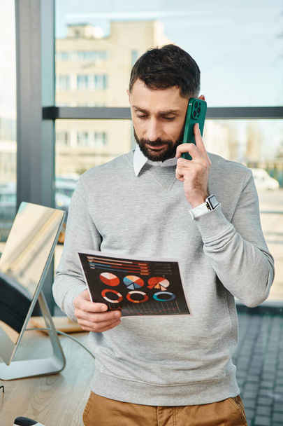 Un uomo in un ambiente aziendale tiene grafici in una mano e parla su un telefono cellulare nell'altra, multitasking per lavoro. - Foto, immagini