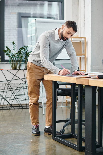 Bir adam dizüstü bilgisayarına odaklanmış, yoğun bir ofiste masa başında duruyor, şirket kültürünün ayrılmaz bir parçası olan bir proje üzerinde çalışıyor.. - Fotoğraf, Görsel