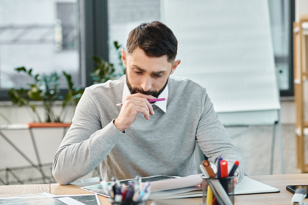 Ein Mann sitzt an einem Schreibtisch, in ein Blatt Papier vertieft, tief in Gedanken, umgeben von der Hektik eines Firmenbüros. - Foto, Bild