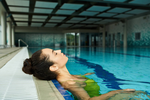 Una joven y hermosa morena en traje de baño flota sin esfuerzo en una piscina cubierta de spa. - Foto, imagen