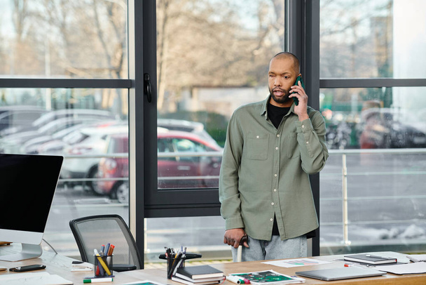 Un hombre con miastenia gravis en traje de negocios hablando en un teléfono celular mientras está parado frente a un escritorio en un entorno de oficina. - Foto, imagen