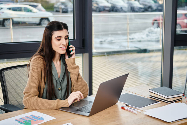 Una donna in un ufficio aziendale siede a un tavolo impegnato in una conversazione su un telefono cellulare. - Foto, immagini