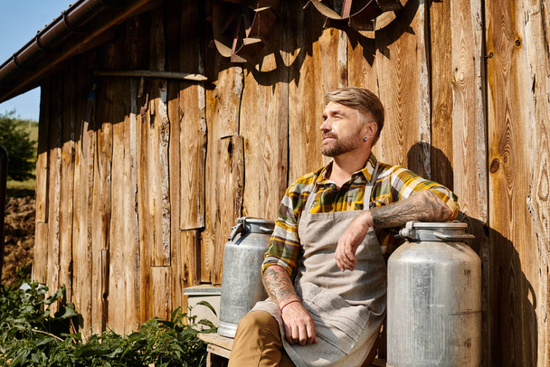 attraente contadino in abbigliamento casual con tatuaggi che tengono l'urna del latte e guardando lontano accanto alla casa - Foto, immagini