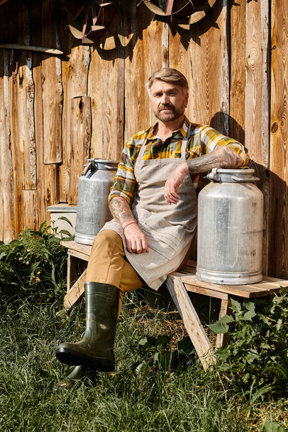 dobře vypadající farmář v ležérní oblečení s tetováním sedí s mléčnými kostkami a dívá se do kamery - Fotografie, Obrázek