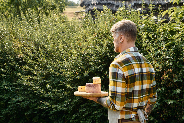 good looking man with tattoos on arms in casual attire holding cheese and looking away while on farm - Fotoğraf, Görsel