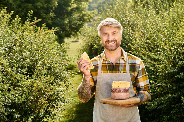 Χαρούμενος όμορφος αγρότης με τατουάζ κρατώντας σπιτικό τυρί στα χέρια του και χαμογελώντας στην κάμερα - Φωτογραφία, εικόνα