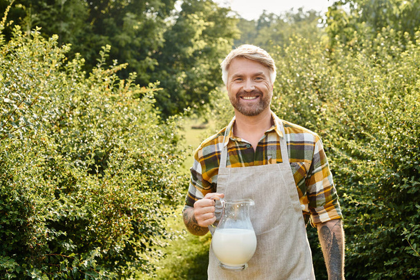 alegre hombre guapo en traje casual con tatuajes sosteniendo frasco de leche fresca y mirando a la cámara - Foto, imagen