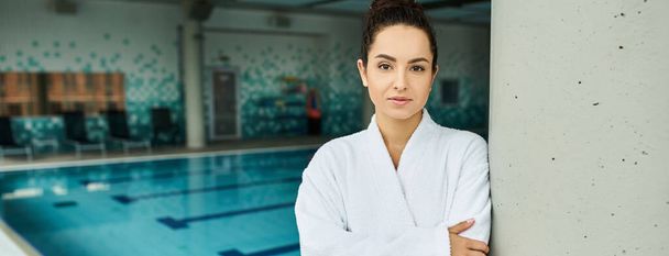 Uma jovem, bela mulher morena de pé elegantemente ao lado de uma piscina interior em um ambiente de spa. - Foto, Imagem