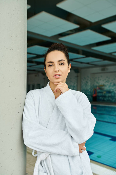 Una mujer joven con albornoz disfruta de un momento tranquilo junto a una piscina cubierta en un entorno de spa. - Foto, imagen