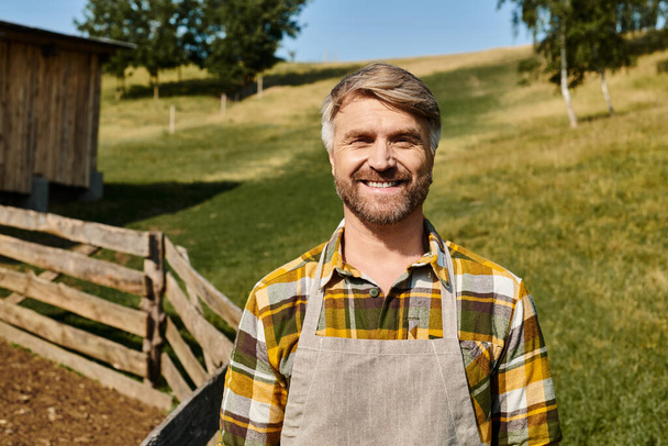 knappe vrolijke man met tatoeages poserend naast hek en mest op boerderij en kijkend naar camera - Foto, afbeelding