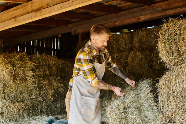 привлекательный преданный человек с бородой и татуировками, работающий с тюками сена на своей ферме - Фото, изображение