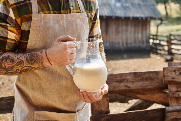 ostříhaný pohled na tvrdě pracující muž s tetováním držící velkou sklenici čerstvého mléka, zatímco na jeho farmě - Fotografie, Obrázek