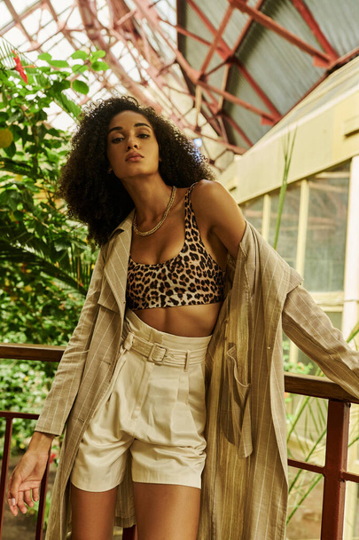 Chic african american woman with curly hair posing in her trendy look in tropical conservatory - Photo, Image