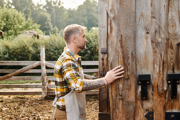ελκυστικός αφοσιωμένος άντρας με γενειάδα και τατουάζ στην αγκαλιά του που κλείνει τις πόρτες του αχυρώνα ενώ είναι στη φάρμα. - Φωτογραφία, εικόνα