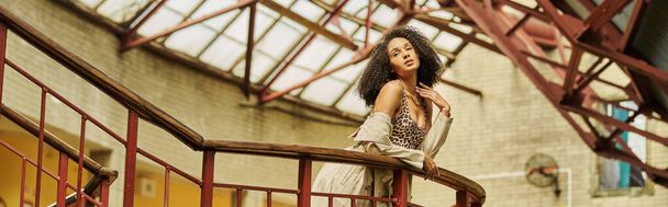 curly chic african american woman in animal print look posing against industrial backdrop, banner - Photo, Image