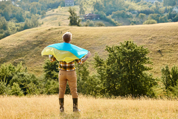 back view of adult Ukrainian farmer with tattoos on arms posing with national flag, scenic landscape - Photo, Image