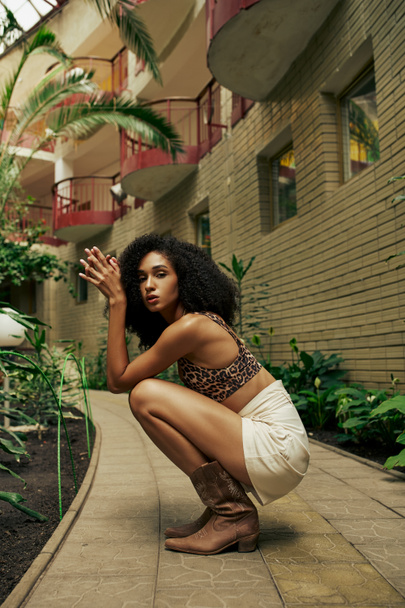 gyönyörű afro-amerikai göndör nő elegáns öltözékben üt egy pózt közel épület zöld növények - Fotó, kép
