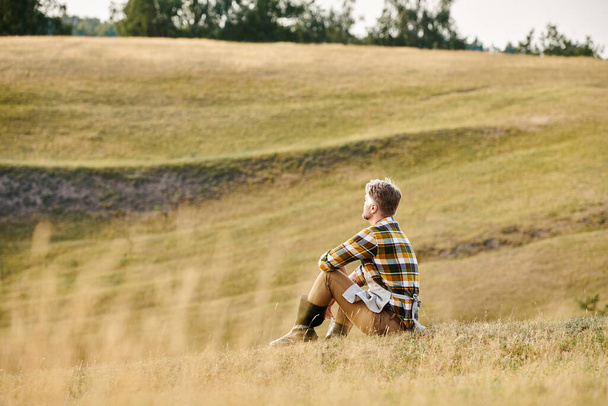 красивый преданный человек с бородой расслабляется на зеленом весеннем поле и смотрит в сторону, современный фермер - Фото, изображение