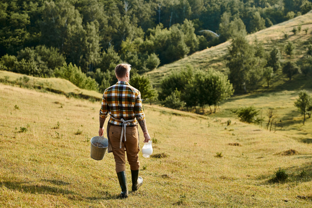 手のバケツとミルクの瓶で歩くタトゥーとハードワーキング現代農家のバックビュー - 写真・画像