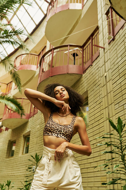 low angle view of black woman in stylish animal print attire posing in atrium with green plants - Photo, Image