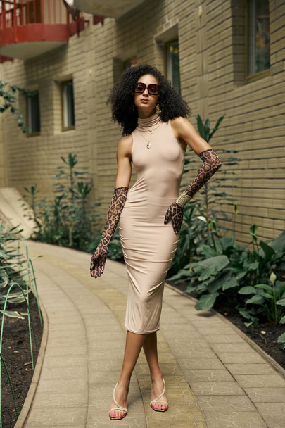 mujer negra joven sofisticada en vestido y gafas de sol posa en guantes de impresión animal en el jardín urbano - Foto, Imagen